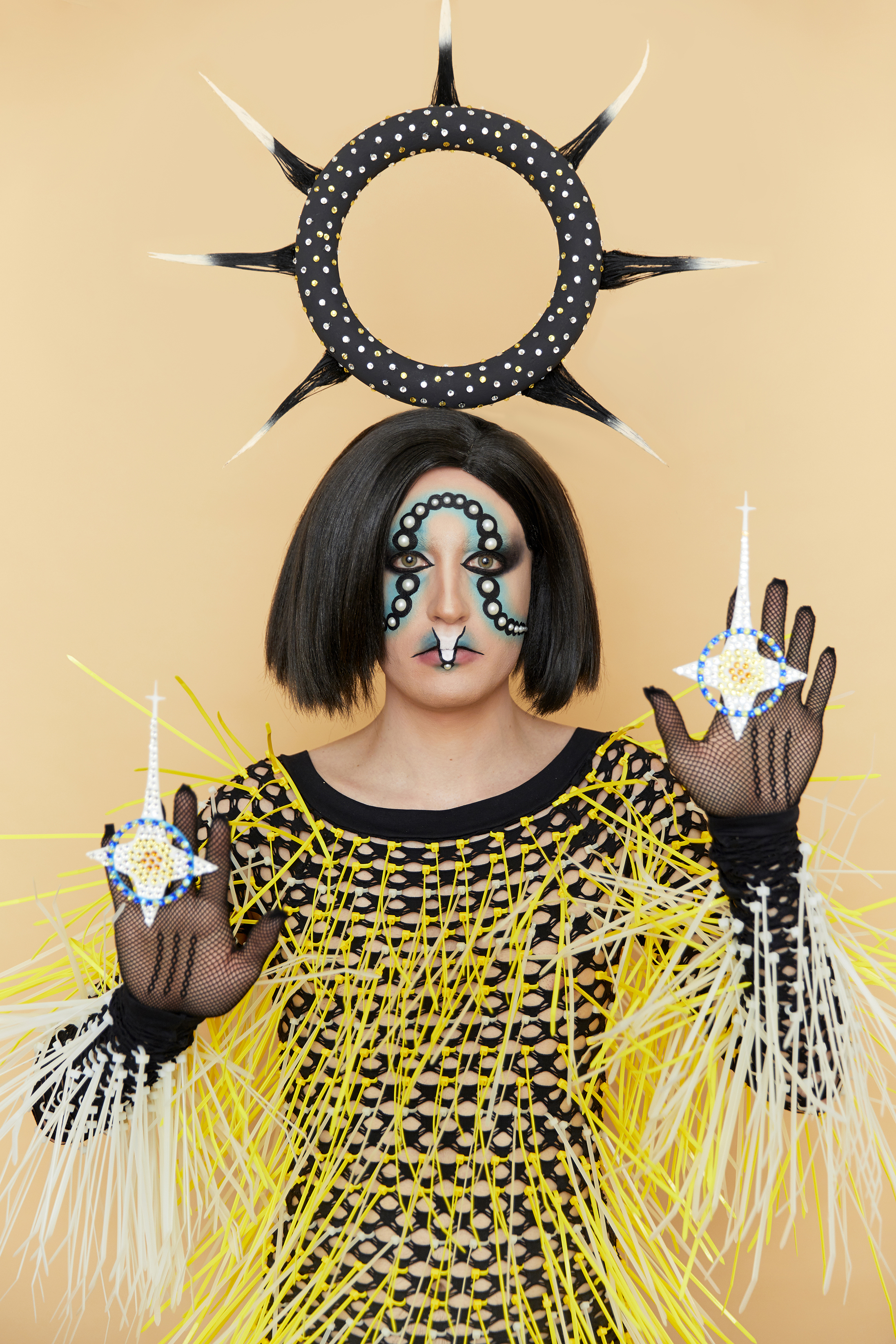 Jesse facing the camera waering a graphic makeup look with an ornate headpiece and black fishnet top covered in yellow and white spikes against a tan background